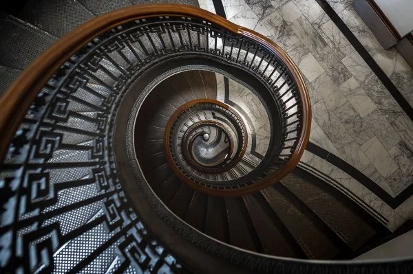Cansu-Meric Gökdemir – Hörzentrum Süderelbe – Ein Blick von oben auf eine elegante Wendeltreppe mit kunstvoll verzierten schwarzen Eisengeländern und Marmorboden.