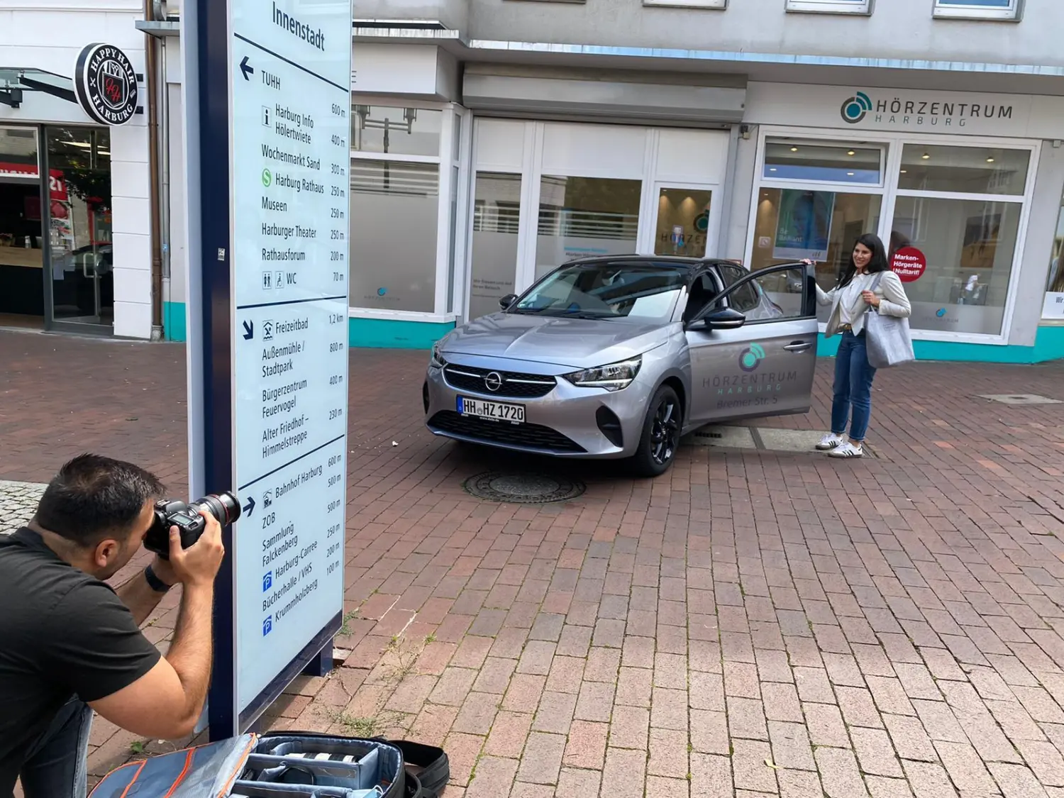 Cansu-Meric Gökdemir – Hörzentrum Süderelbe – Ein Fotograf macht ein Foto von einem silbernen Opel Corsa, der neben einer Person an der offenen Autotür auf einer gepflasterten Straße parkt.