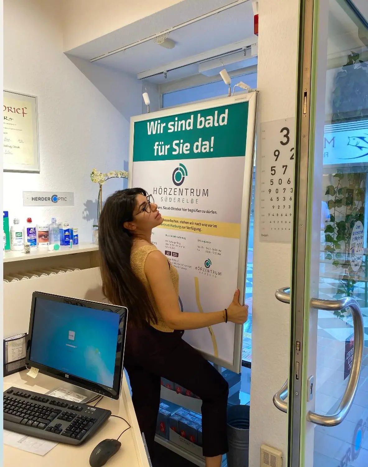 Cansu-Meric Gökdemir – Hörzentrum Süderelbe – Eine Person sitzt an einem Schreibtisch in einem Büro oder Beratungsraum vor einer gemusterten Wand und einem Schild an der Wand im Hintergrund.